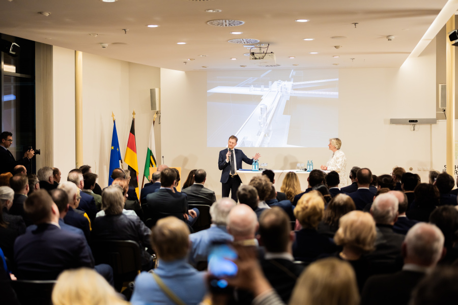 Männlich gelesene Person spricht in ein Mikrofon vor einer Gruppe von Menschen.