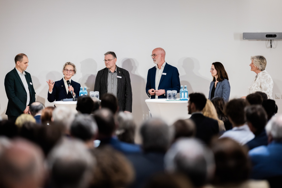 Eine Gruppe von Menschen steht vor einem Publikum. Eine weiblich gelesene Person hält ein Mikrofon in der Hand und spricht hinein.
