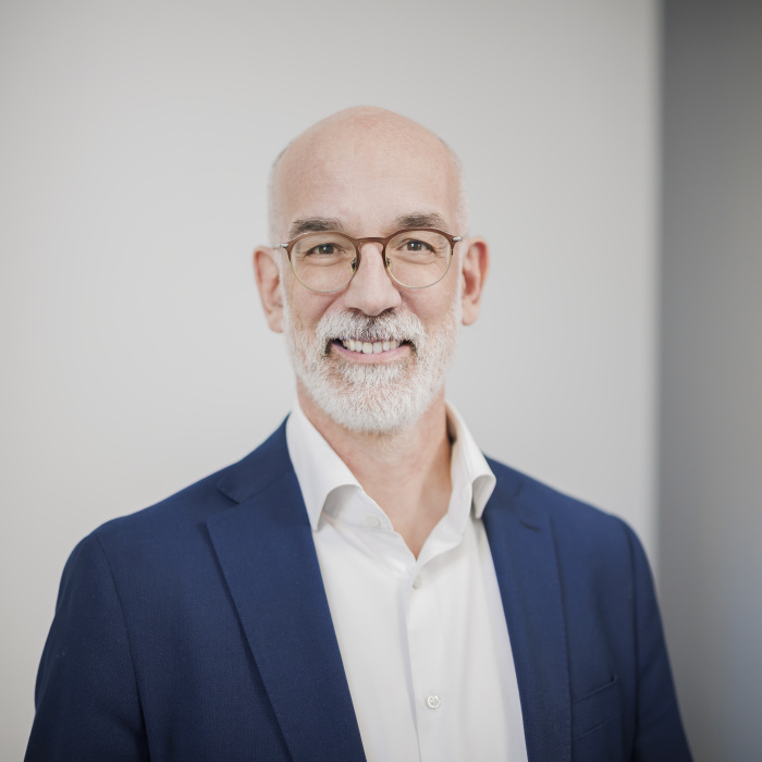 Männlich gelesene Person mit Brille blickt lächelnd in die Kamera.