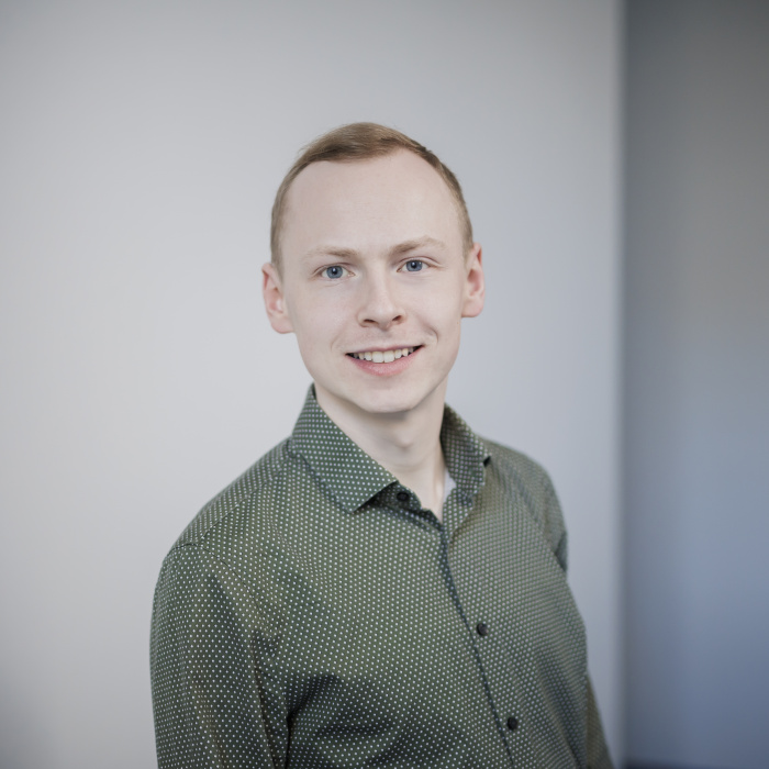 Männlich gelesene Person mit kurzen hellen Haaren und grünem Hemd blickt lächelnd in die Kamera.